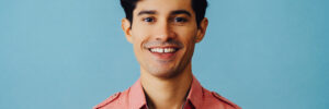 Headshot hispanic latino man black hair smiling handsome young adult wearing pink shirt over blue background looking at camera studio shot