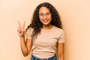 Young hispanic woman isolated on beige background showing victory sign and smiling broadly.