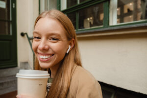 Portrait of young smiling woman with blonde hair listening music with earphones and drinking coffee while sitting outdoors