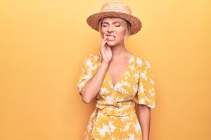Beautiful blonde woman on vacation wearing summer hat and dress over yellow background touching mouth with hand with painful expression because of toothache or dental illness on teeth.